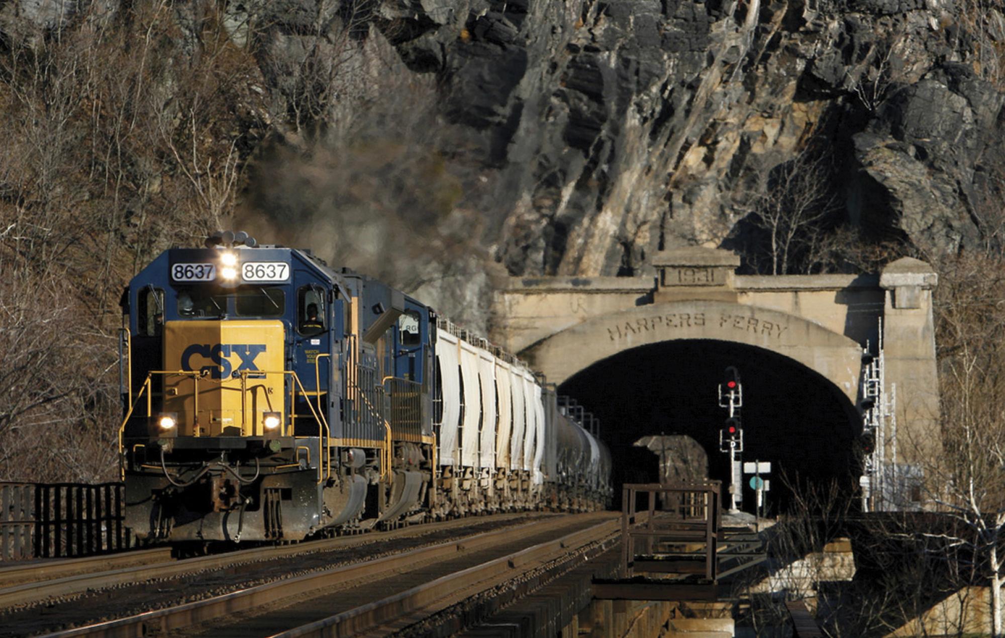 A yellow and blue CSX train on a track emerges from a tunnel that cuts through a mountain